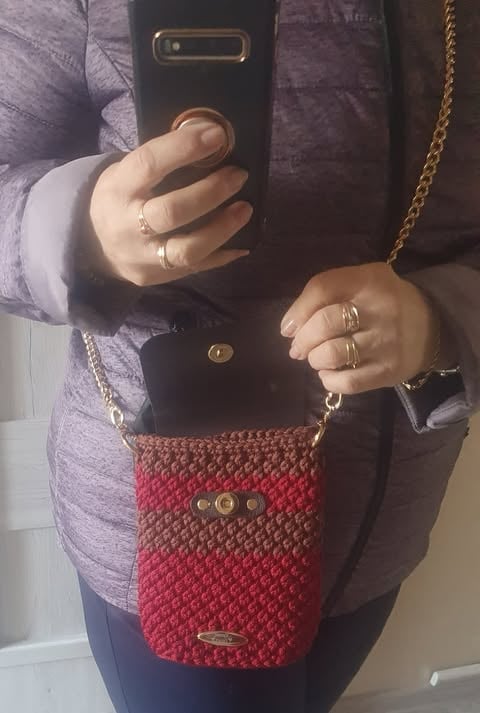 Person holding a black phone and red crossbody bag with gold chain strap, wearing multiple gold rings