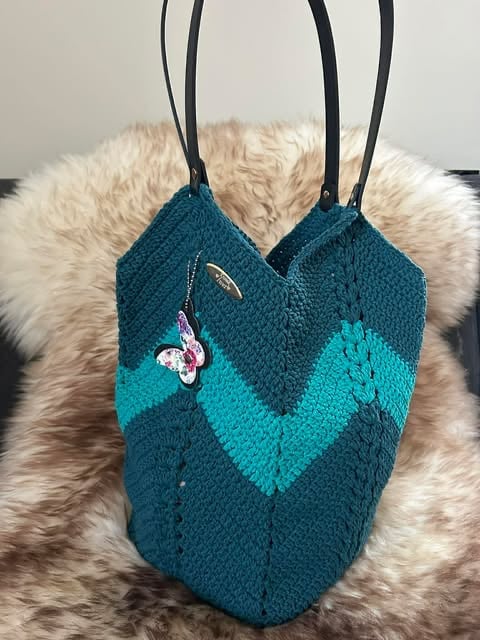 Teal knitted handbag with turquoise color-blocked pattern, beaded butterfly motif, and black straps on fuzzy background