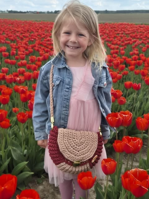 Young girl smiling in a red tulip field wearing a denim jacket, pink dress, and woven crossbody bag