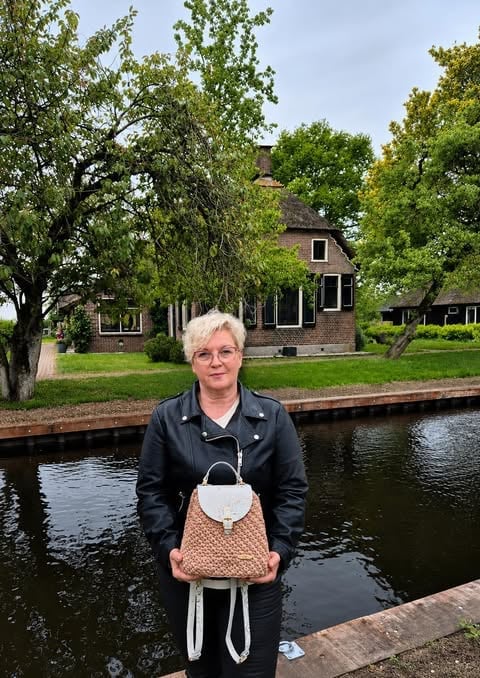 Woman with short white hair wearing black jacket standing by canal with brick house and trees in background