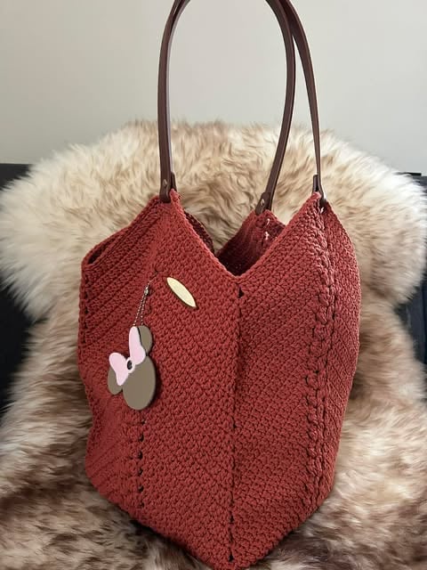 Burgundy knit tote bag with brown leather handles and decorative cutouts, displayed on a cream-colored fur backdrop