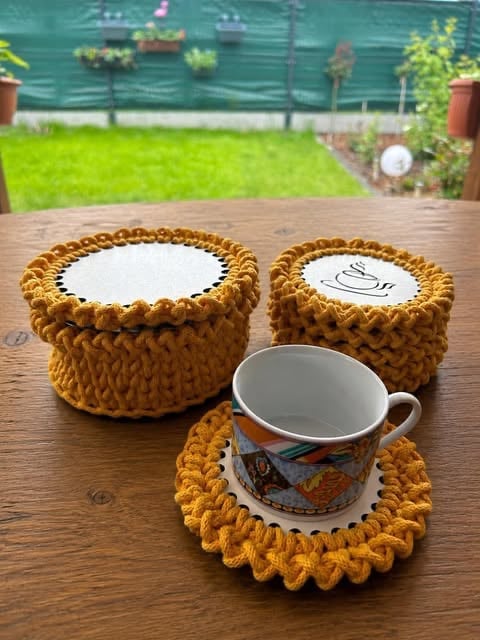 Three knitted mustard-yellow coasters on wooden table outdoors, one holding a decorative mug with garden visible in background