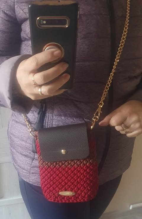 Person holding black smartphone with knitted red crossbody bag featuring gold chain strap and dark leather flap