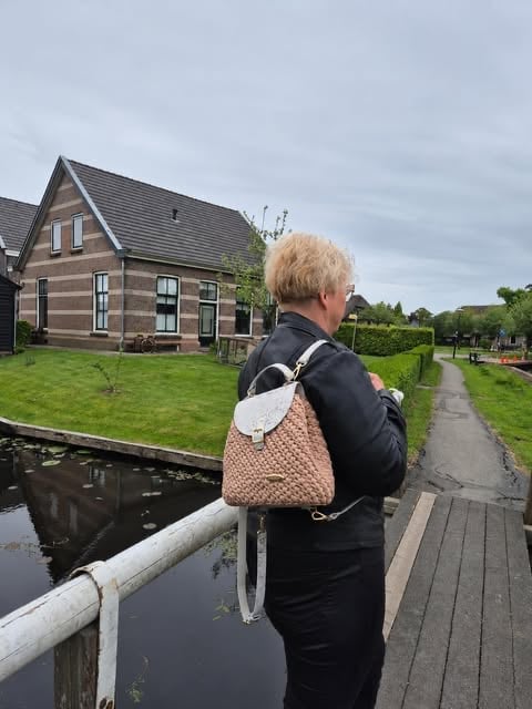 Person with blonde hair and black jacket wearing a beige backpack stands on a wooden bridge overlooking a traditional Dutch house with green lawn and canal