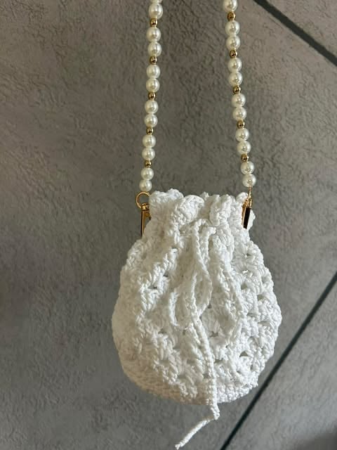 White crocheted drawstring pouch pendant hanging from pearl and gold beaded chain necklace against neutral background