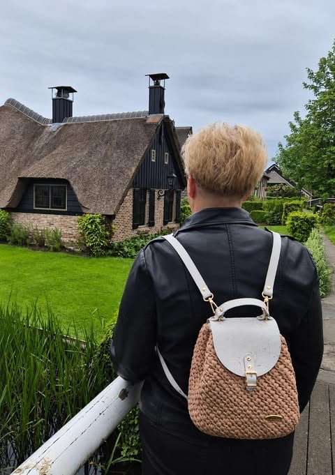 Person with blonde hair wearing black jacket and tan backpack viewing traditional thatched-roof cottage with wooden chimneys