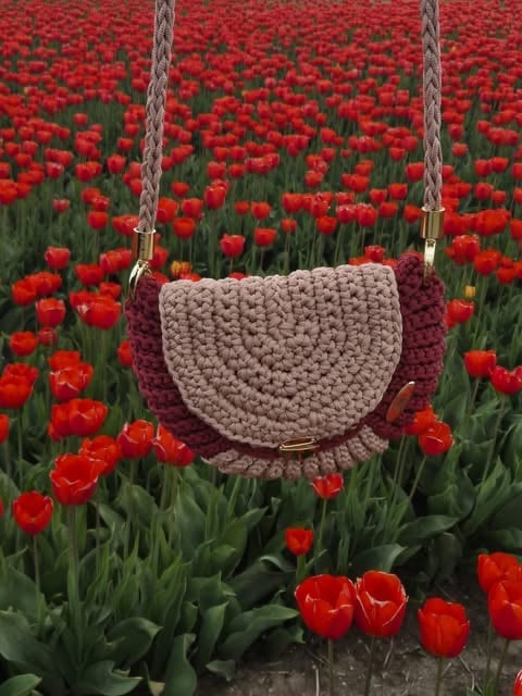 Crocheted shoulder bag in beige and burgundy hanging in a field of blooming red tulips