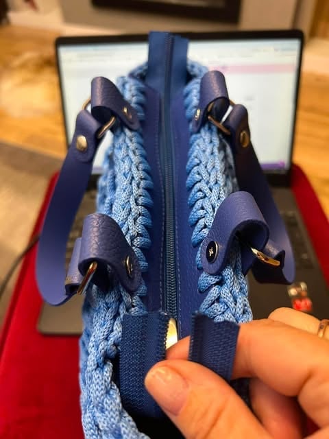 Close-up of a blue knitted pouch with navy zipper and leather handles held open with fingers