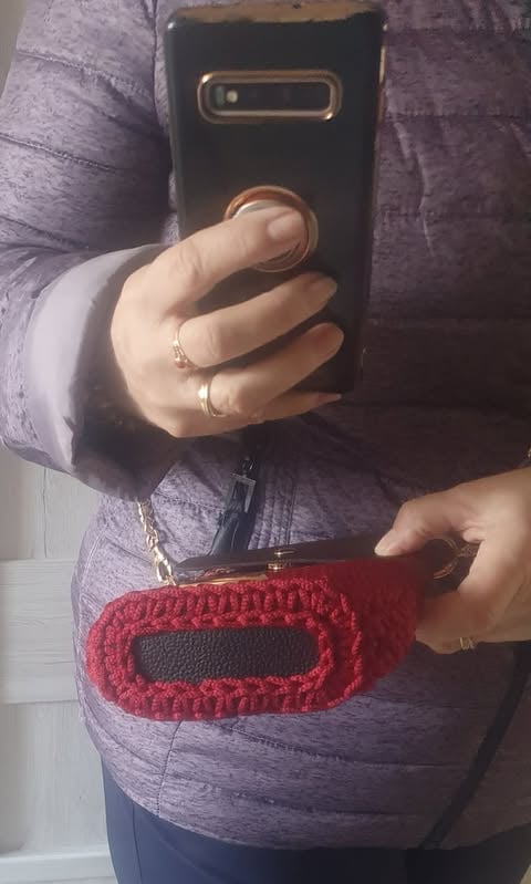 Person holding a black smartphone with gold accents, wearing rings and a red knitted phone pouch around their neck