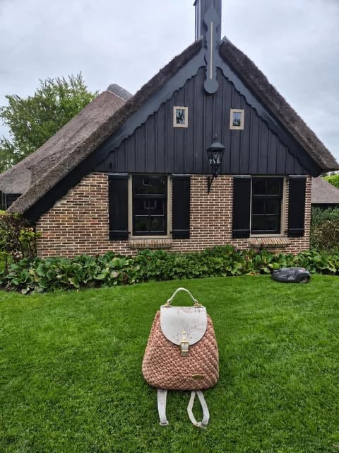 A brown knitted backpack sitting on green grass in front of a brick cottage with a thatched roof