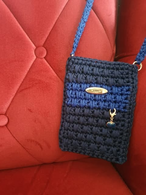 Small knitted crossbody bag in navy and blue with blue strap, displayed on red quilted furniture