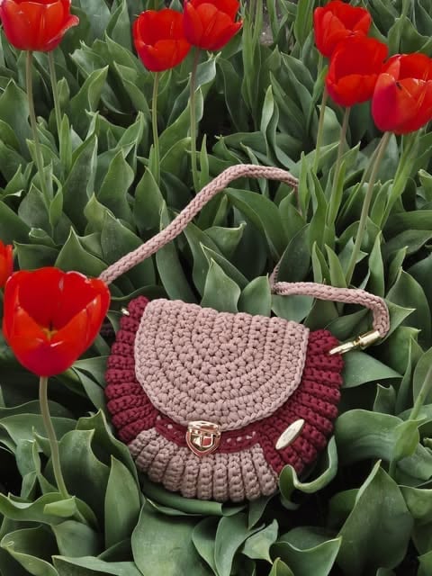 A hand-crocheted pink and burgundy shoulder bag sitting among bright red tulips and green foliage